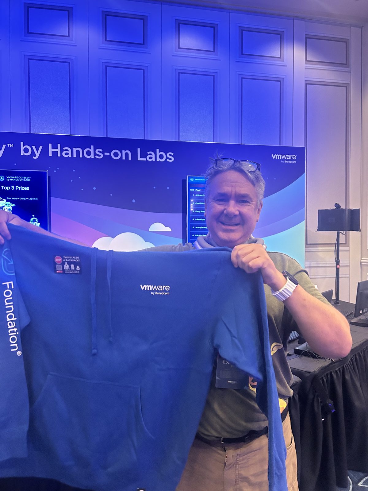 Robert Greenlee holding up a blue VMware hoodie at the Odyssey by Hands-on Labs stand, leaderboard visible behind him