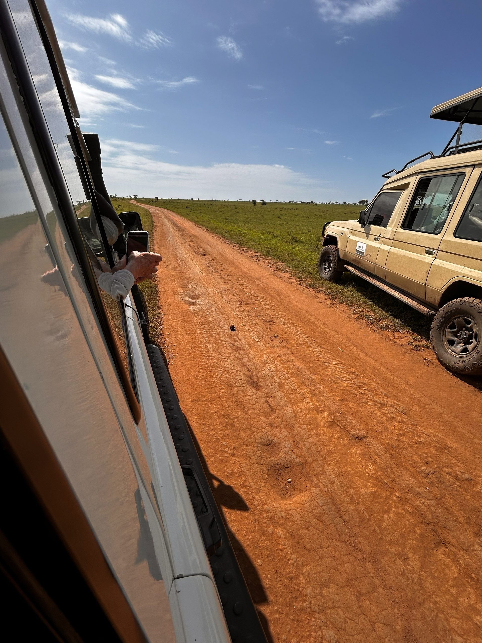 Two Thomson Safari Land Cruisers on the red dirt road across the Serengeti plain — a hand reaching out the window