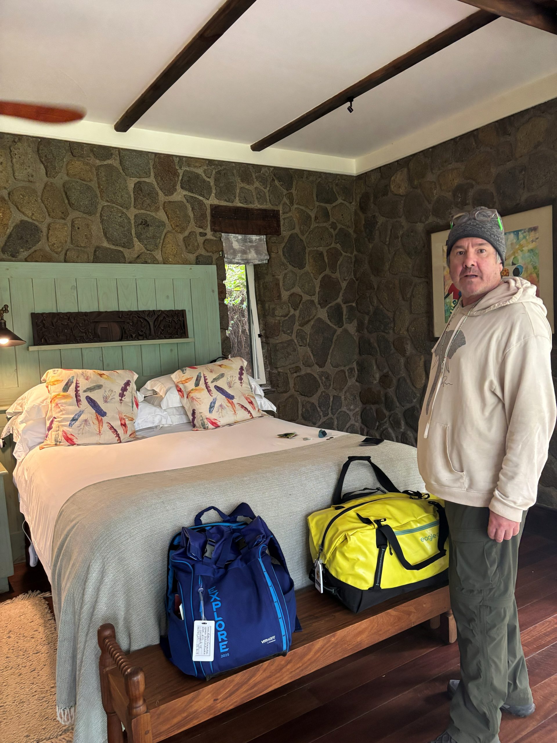 Robert standing beside the bed in the Gibb's Farm Tloma cottage — rough stone walls, sage-green headboard with carved panel, feather-print pillows, timber beams overhead, safari bags on the bench