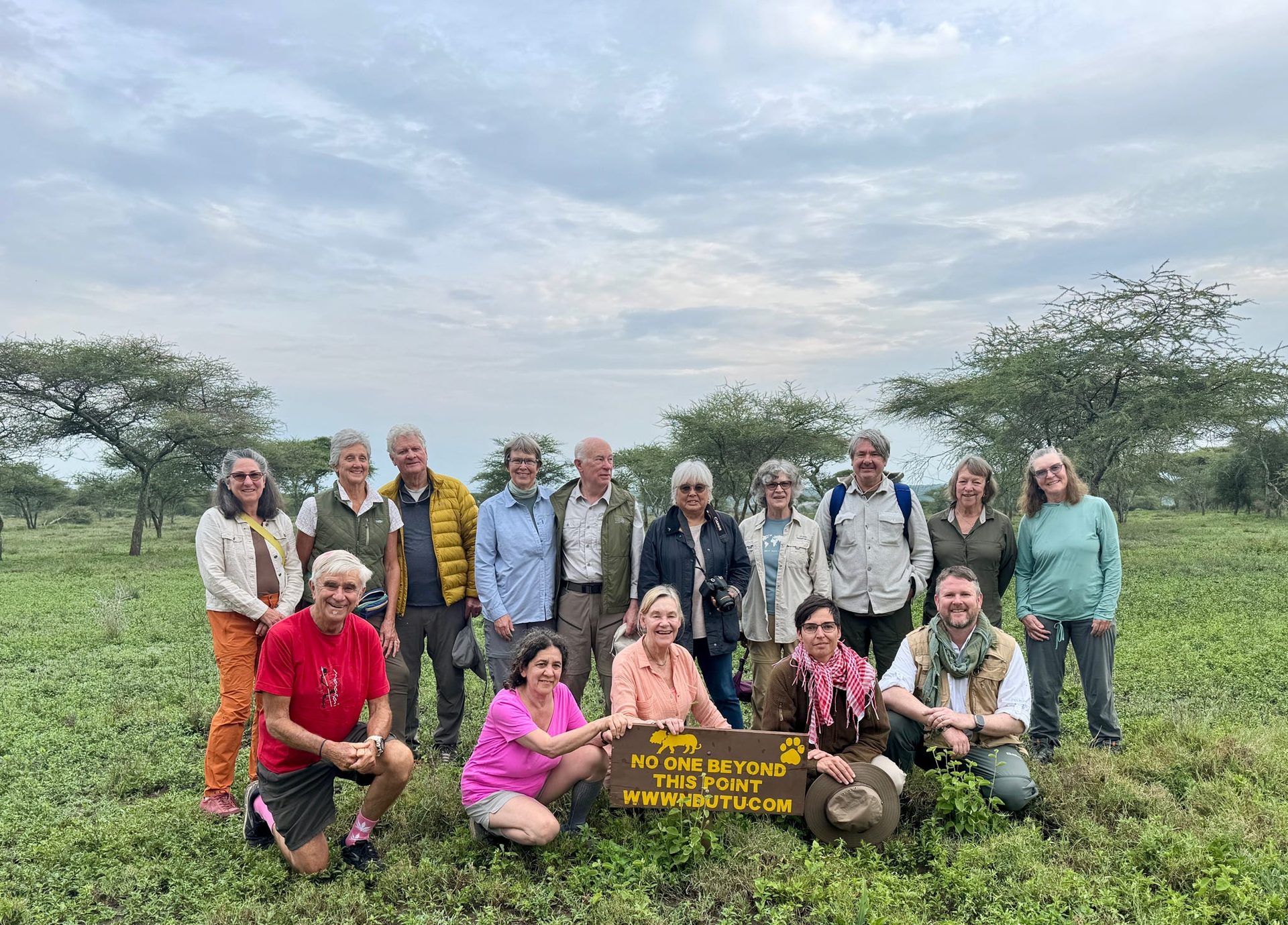 The full group photo at the Ndutu Lodge boundary sign — No One Beyond This Point, wwwndutu.com — everyone grinning, the acacia woodland behind