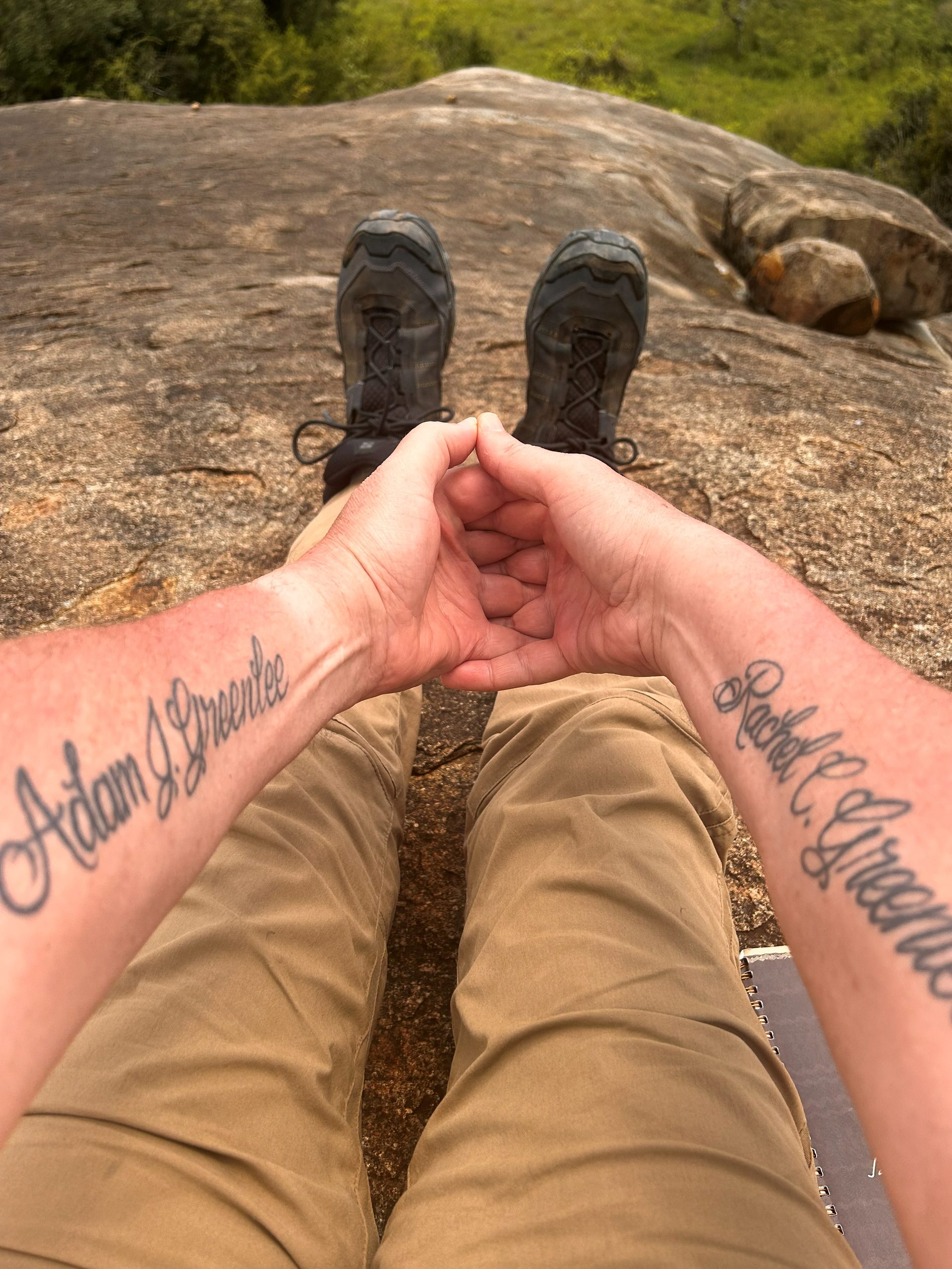 Robert sitting cross-legged on the Gong Rock granite surface, hands in a meditation gesture, forearms bearing tattoos reading Adam J. Greenlee and Rachel C. Greenlee