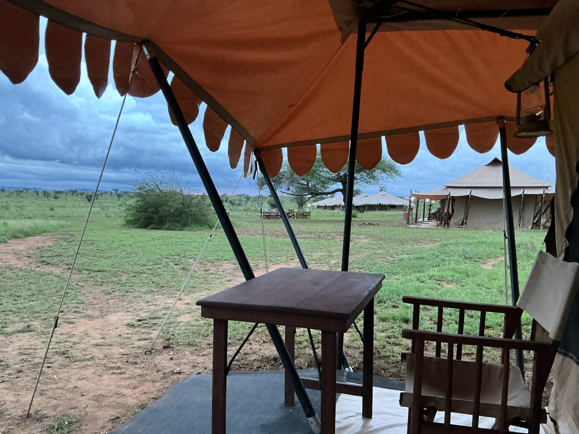 View from the Nyumba tent porch — orange canvas awning, wooden table and chair, green Serengeti plain stretching to the thatched dining tent