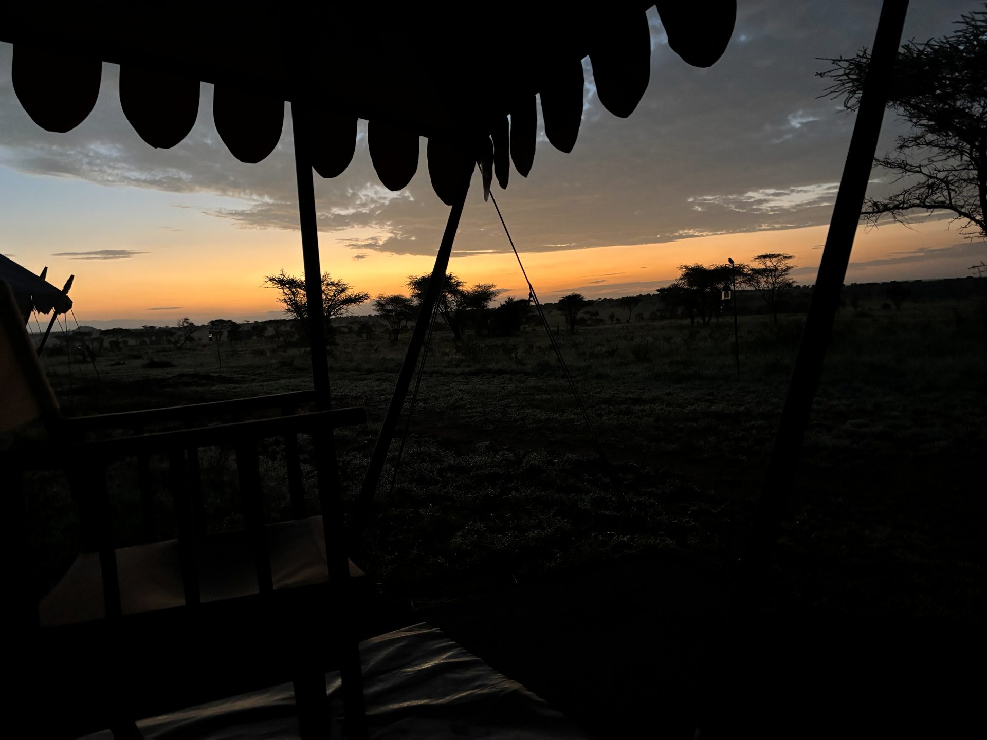 Sunset from the tent porch at the Nyumba camp — scalloped canvas awning silhouetted against a deep orange sky, acacia trees darkening on the plain below