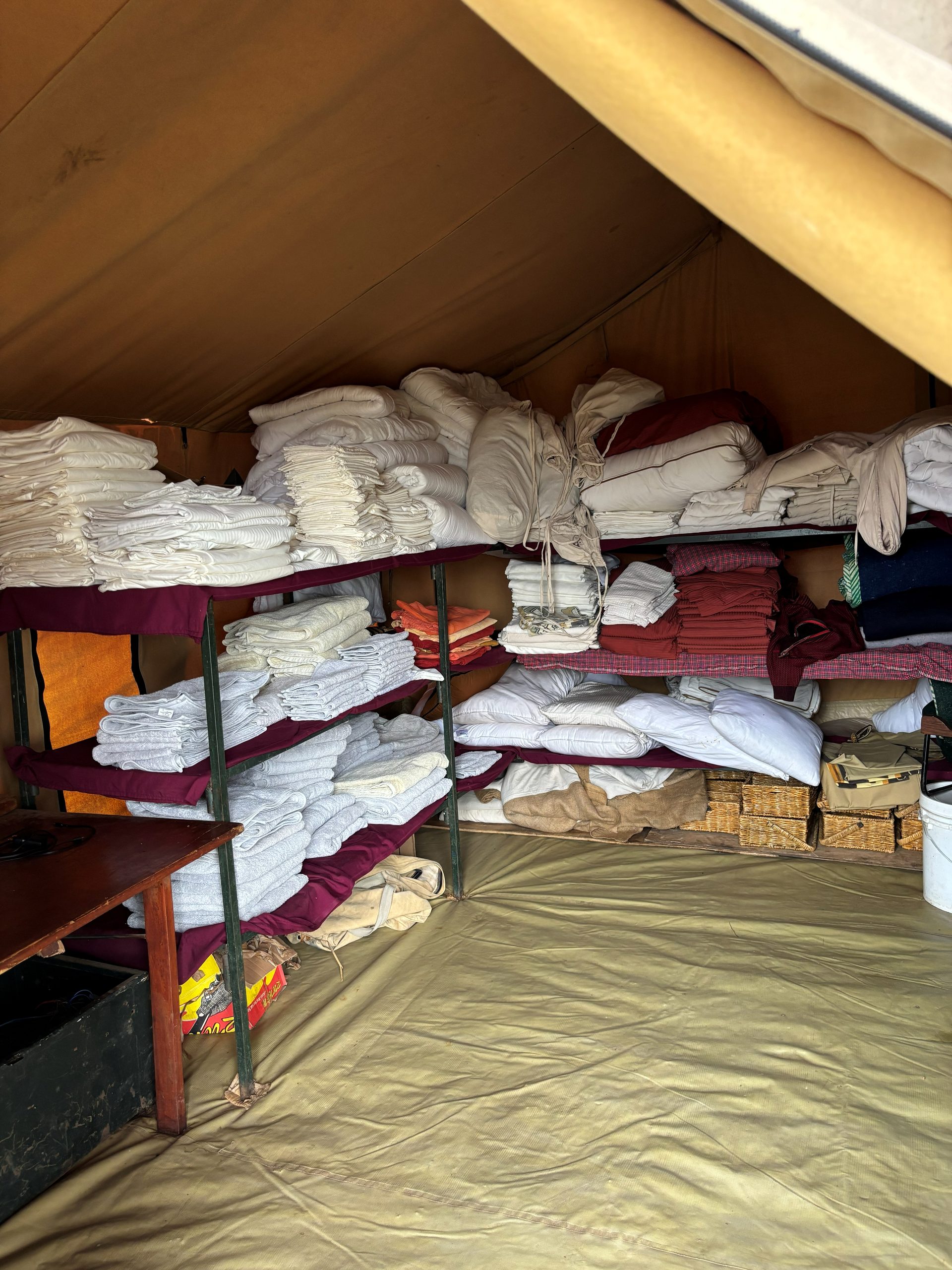 The camp linen tent — floor-to-ceiling shelving stacked with neatly folded white sheets, pillowcases, burgundy blankets, and grey towels