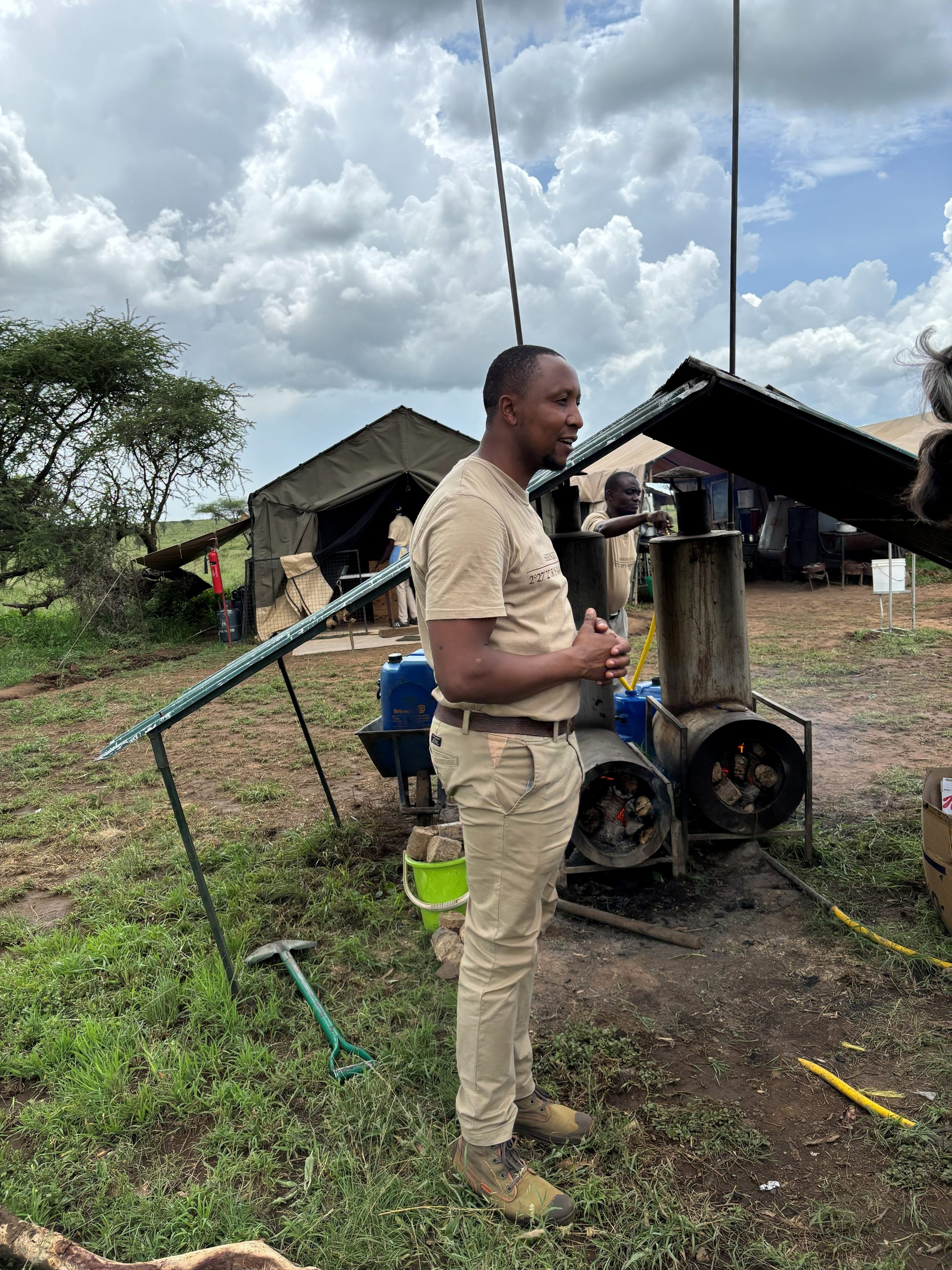 A camp staff member explaining the wood-fired water heating boiler outside — a solar panel visible to the left, the kitchen tent behind